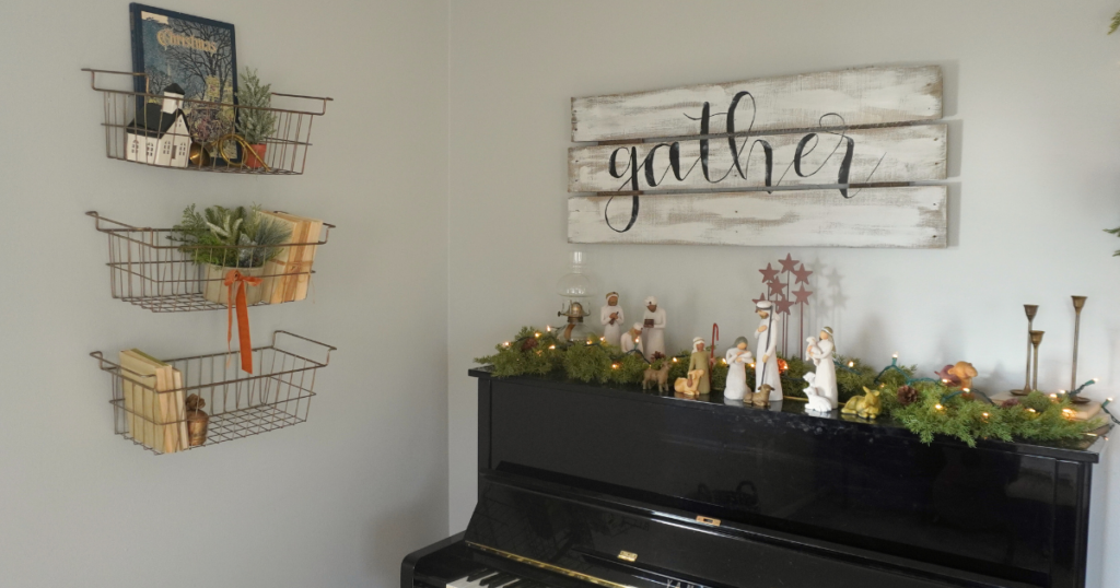 Piano with nativity on top with garland, lights, candles and lamp. Three wire baskets on wall with greenery, bells ribbons and other decor.