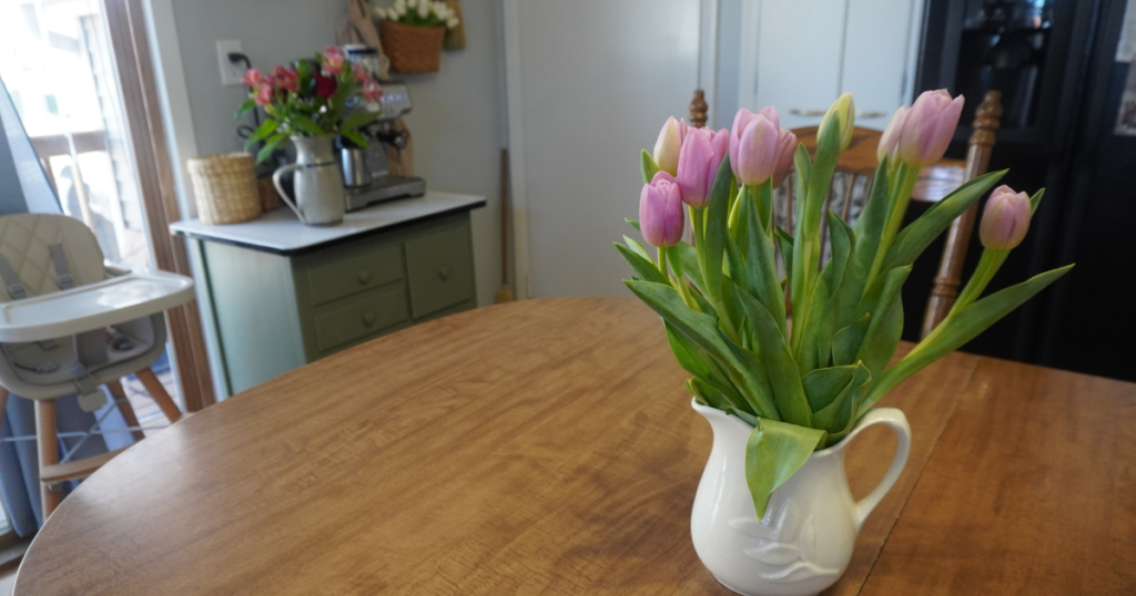 Pink tulips on a table with green stand and pitcher of other flowers and accordian rack with basket of tulips.