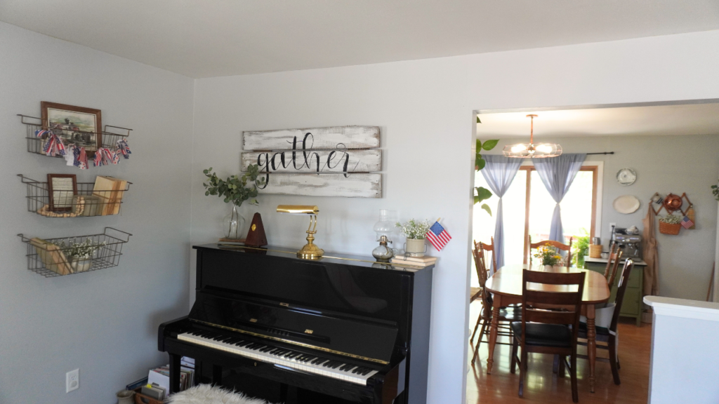 black piano with decor on top and wire baskets on wall with patriotic banner and books view of kitchen in background