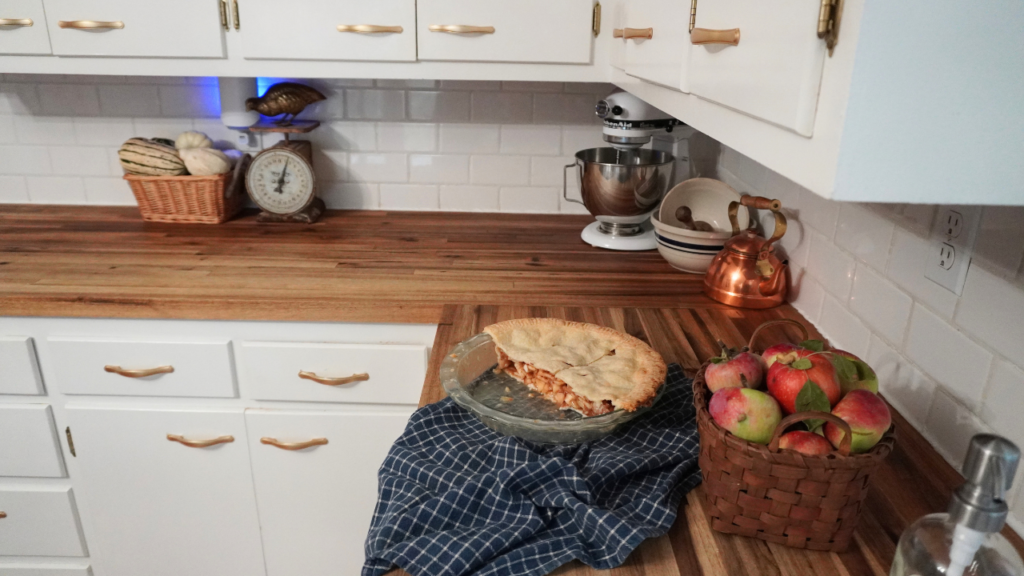 Half eaten apple pie with basket of apples next to it. Copper tea kettle and bowls on coutner with pumpkins in basket.