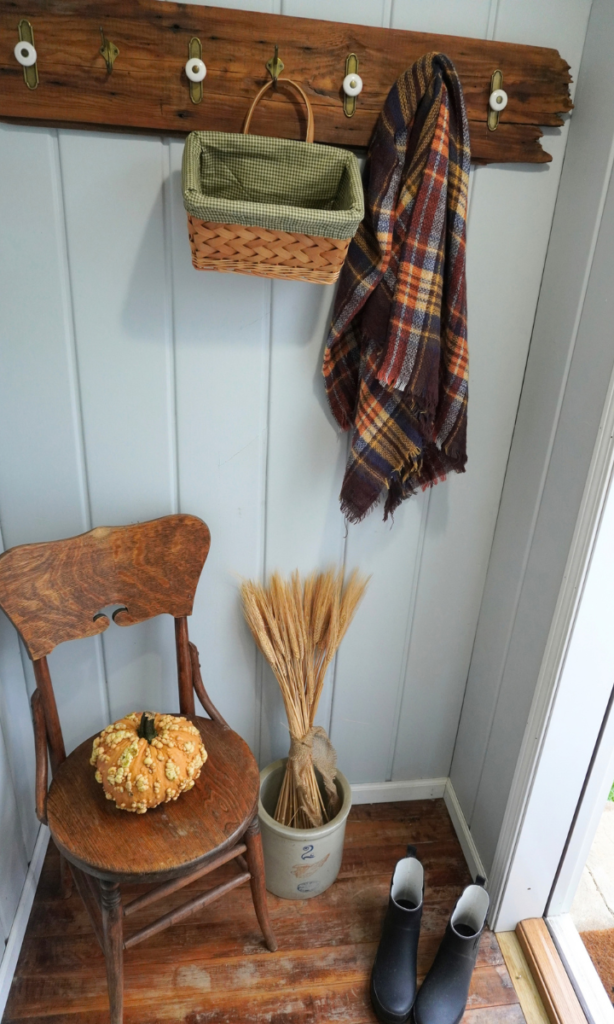 wood chair with pumpkin on it, wheat stems in crock and boots on floor. basket and fall blanket on coat hook.