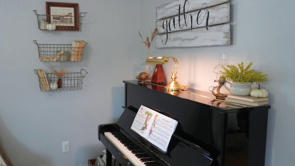 Black piano with faux yellow flowers and pumpkin on it. Wire baskets on wall with wheat stems, books, fall art.